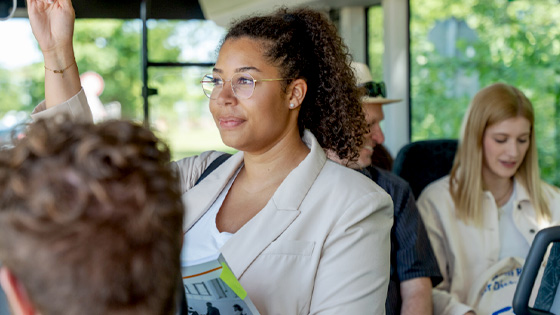 Eine junge Frau mit lockigen Haaren und Brille steht lächelnd im Bus und hält sich an einer Haltestange fest