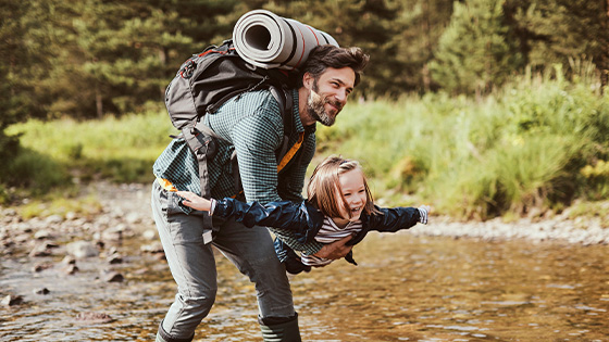 Ein Mann mit großem Rucksack trägt ein lachendes kleines Mädchen spielerisch, die Arme ausgestreckt, durch einen flachen Bachlauf in der Natur