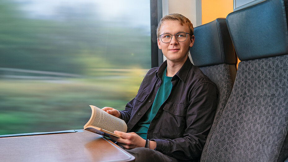 A young man with glasses sits by the window on a train and reads a book