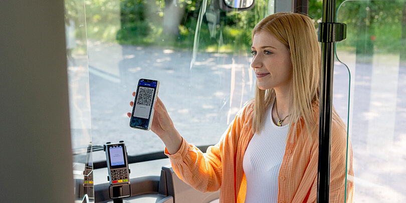 Eine junge Frau in einer orangefarbenen Strickjacke zeigt ihren mobilen Fahrschein (QR-Code) auf dem Smartphone dem Busfahrer oder Kontrolleur