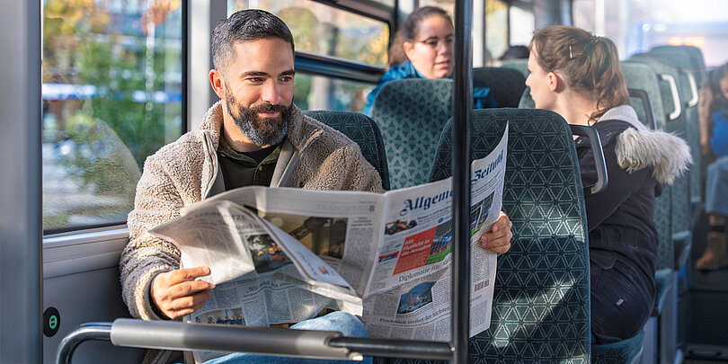 Mann mit Bart in einem hellbraunen Fleece-Jacket sitzt in einem Bus und liest eine Zeitung, während andere Fahrgäste auf den Sitzen dahinter sitzen