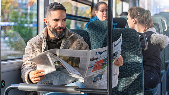 Mann mit Bart in einem hellbraunen Fleece-Jacket sitzt in einem Bus und liest eine Zeitung, während andere Fahrgäste auf den Sitzen dahinter sitzen