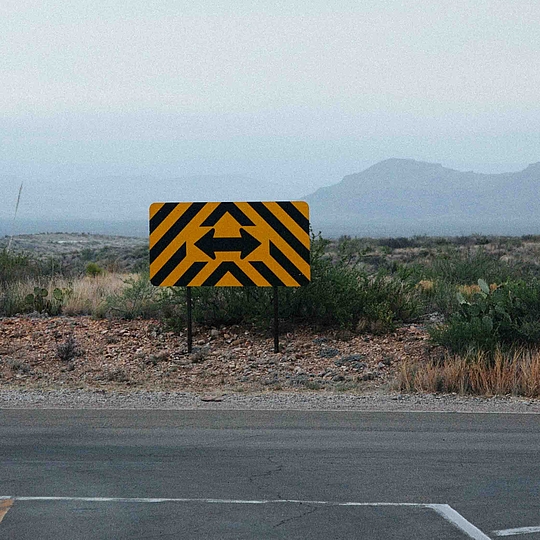 Gelbes Warnschild mit schwarzen Pfeilen auf ländlicher Straße
