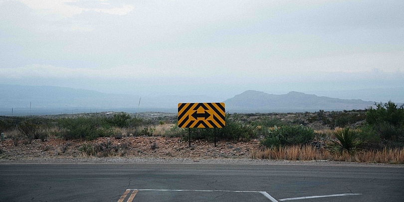 Gelbes Warnschild mit schwarzen Pfeilen auf ländlicher Straße