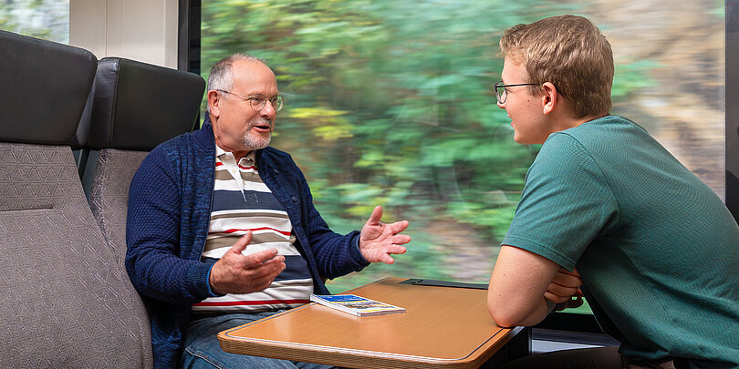 2 Männer sitzen im Zug an einem Tisch gegenüber: Ein älterer und ein jüngerer Mann.