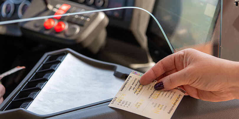 Nahaufnahme: Die Hand eines Fahrgastes reicht ein Papierticket an den Busfahrer über den Fahrkartenschlitz
