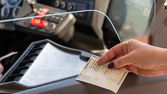 Nahaufnahme: Die Hand eines Fahrgastes reicht ein Papierticket an den Busfahrer über den Fahrkartenschlitz