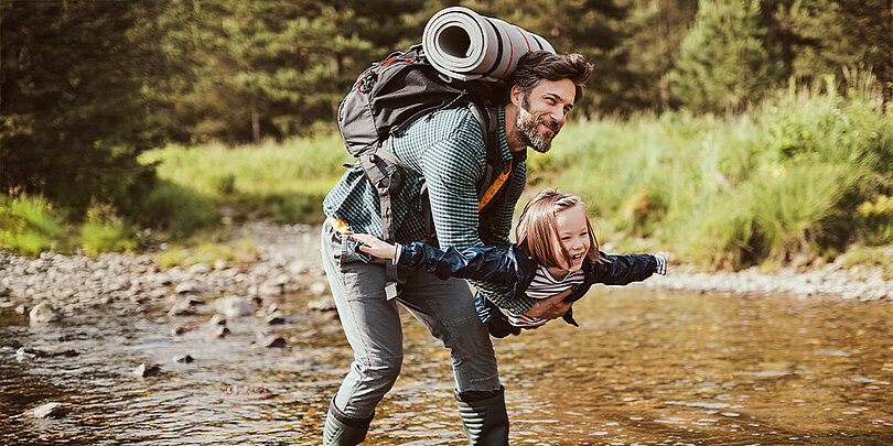 Ein Mann mit großem Rucksack trägt ein lachendes kleines Mädchen spielerisch, die Arme ausgestreckt, durch einen flachen Bachlauf in der Natur