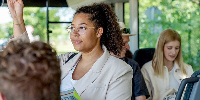 Eine junge Frau mit lockigen Haaren und Brille steht lächelnd im Bus und hält sich an einer Haltestange fest