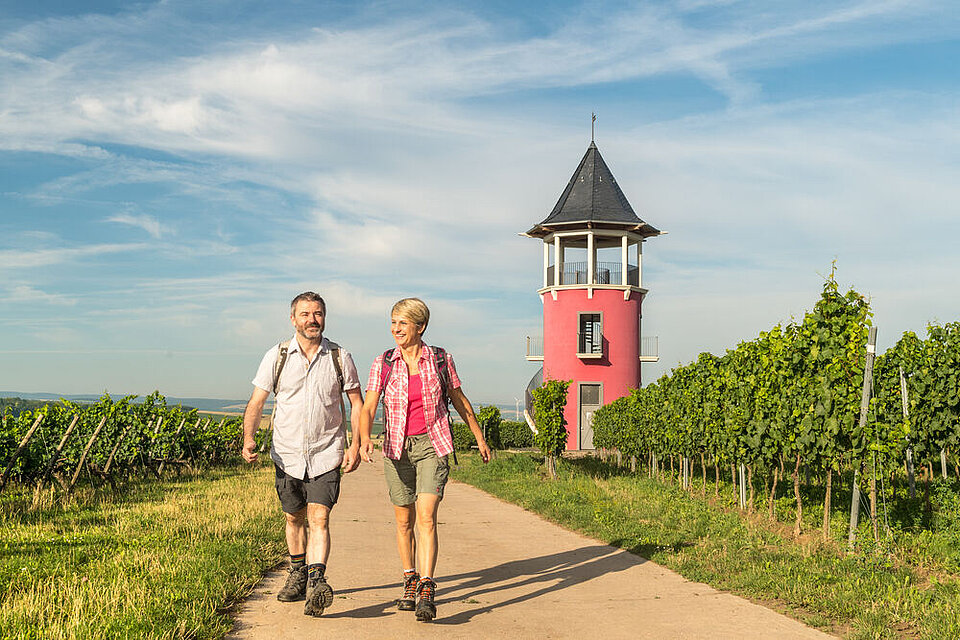 Zwei lächelnde Wanderer, ein Mann und eine Frau, gehen auf einem Weg durch einen sonnigen Weinberg. Im Hintergrund steht ein kleiner, markanter roter Turm