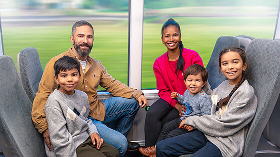 Eine Familie sitzt in einem Zug sich gegenüber und schaut in die Kamera.