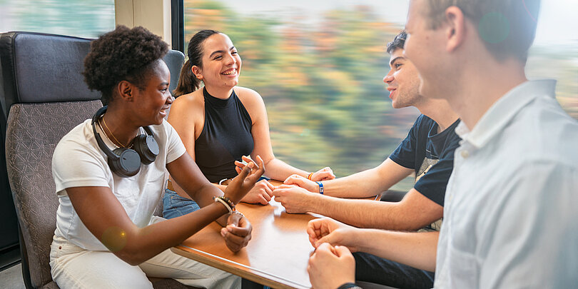Vier junge Menschen sitzen lachend an einem Tisch in einem fahrenden Zugabteil