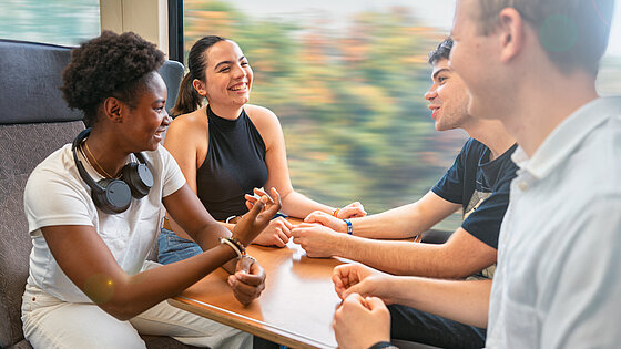 Vier junge Menschen sitzen lachend an einem Tisch in einem fahrenden Zugabteil