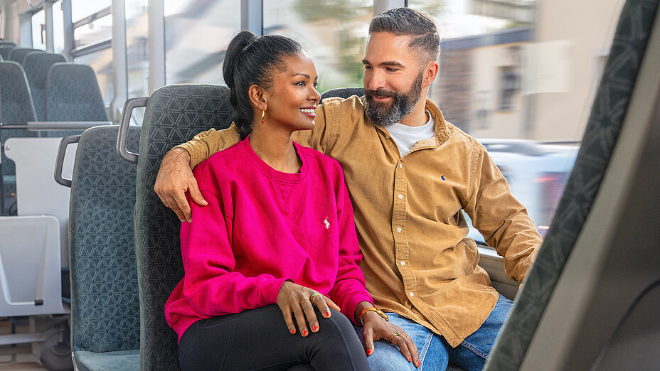 A couple is sitting together on the bus. The man has put his arm around the woman, and they are smiling at each other