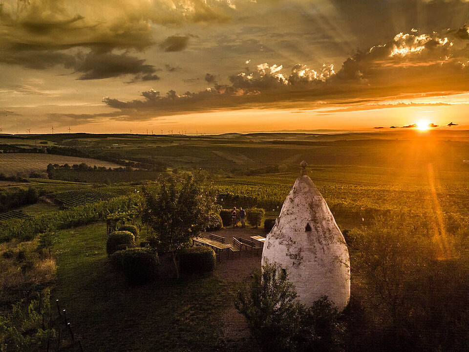 Eine Drohnenaufnahme einer kegelförmigen, weißen Hütte und einer Sitzgruppe in einem Weinberg bei Sonnenuntergang. Am Horizont sind Windräder zu sehen