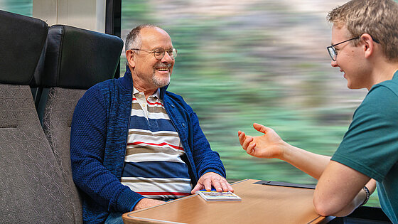 Zwei Männer, ein älterer und ein jüngerer, unterhalten sich lachend in einem Zugabteil mit Tisch