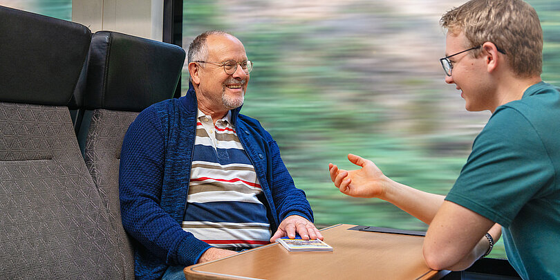 Zwei Männer, ein älterer und ein jüngerer, unterhalten sich lachend in einem Zugabteil mit Tisch