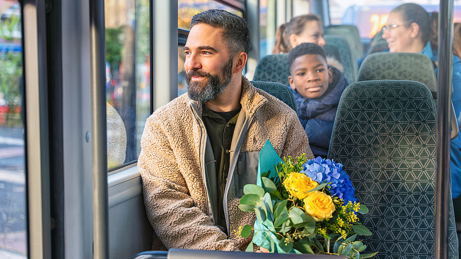 A bearded man in a light jacket sits by the bus window and holds a bouquet of yellow and blue flowers