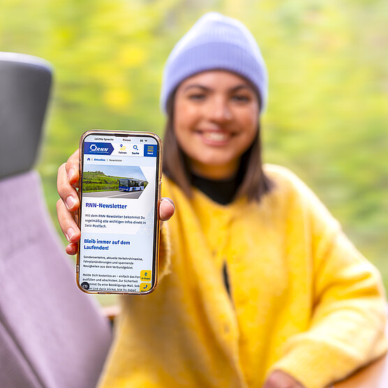Newsletter-Anmeldung Einer lächelnden Frau, die in einem öffentlichen Verkehrsmittel steht und ein Smartphone mit einer geöffneten mobilen Webseite hält. Auf der weißen Überlagerung steht der Text: "Newsletter-Anmeldung"