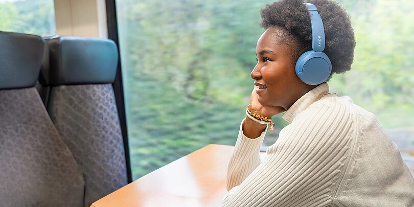 Eine junge Frau sitzt in der Bahn an einem Tisch und hat einen Kopfhörer auf.