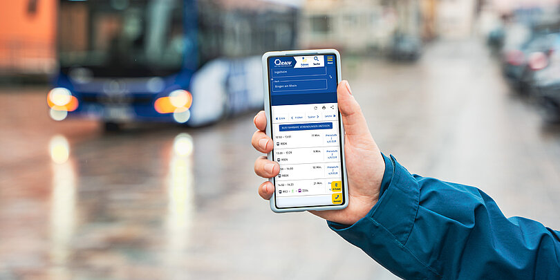 Eine Hand hält ein Mobiltelefon mit der RNN Fahrplanauskunft. Im Hintergrund ist ein Bus zu sehen..
