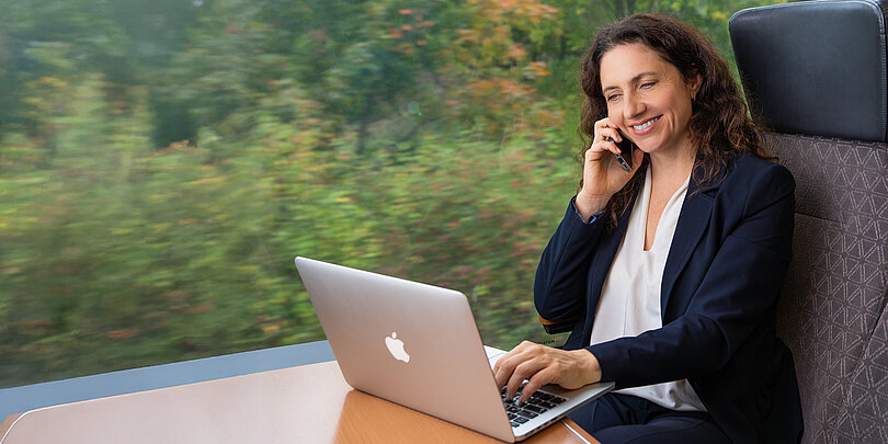 Woman on the train talking on the phone with a laptop in front of her.