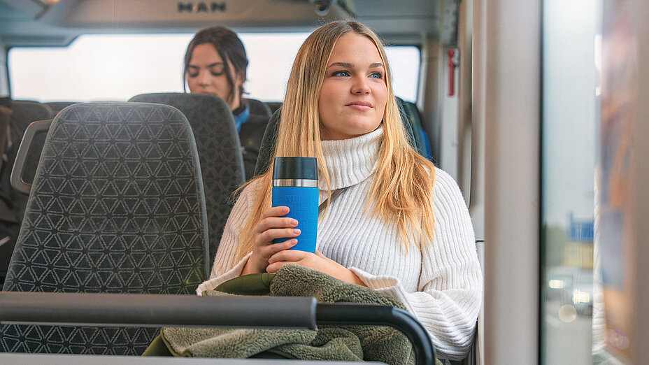 A very beautiful young blonde woman in a white pullover sits by the window on the bus, looks into the distance and holds a thermos with a tea